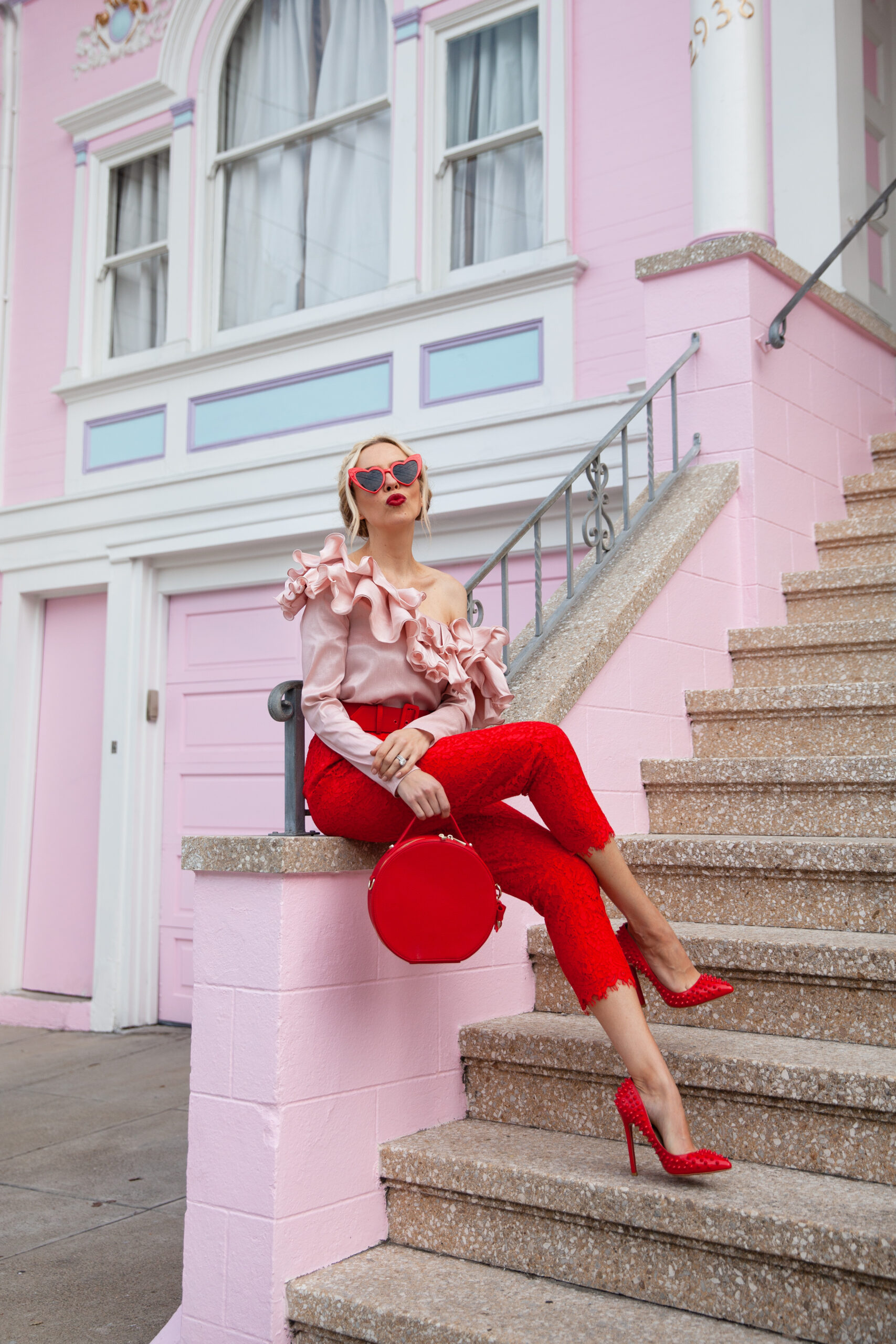 Valentines Day self care featured by top US life and style blog, Lombard and Fifth: image of a blonde woman wearing DressLily heart sunglasses, Roy Rogers red pants, a pink one shoulder ruffle blouse, Michka jeweled red pumps