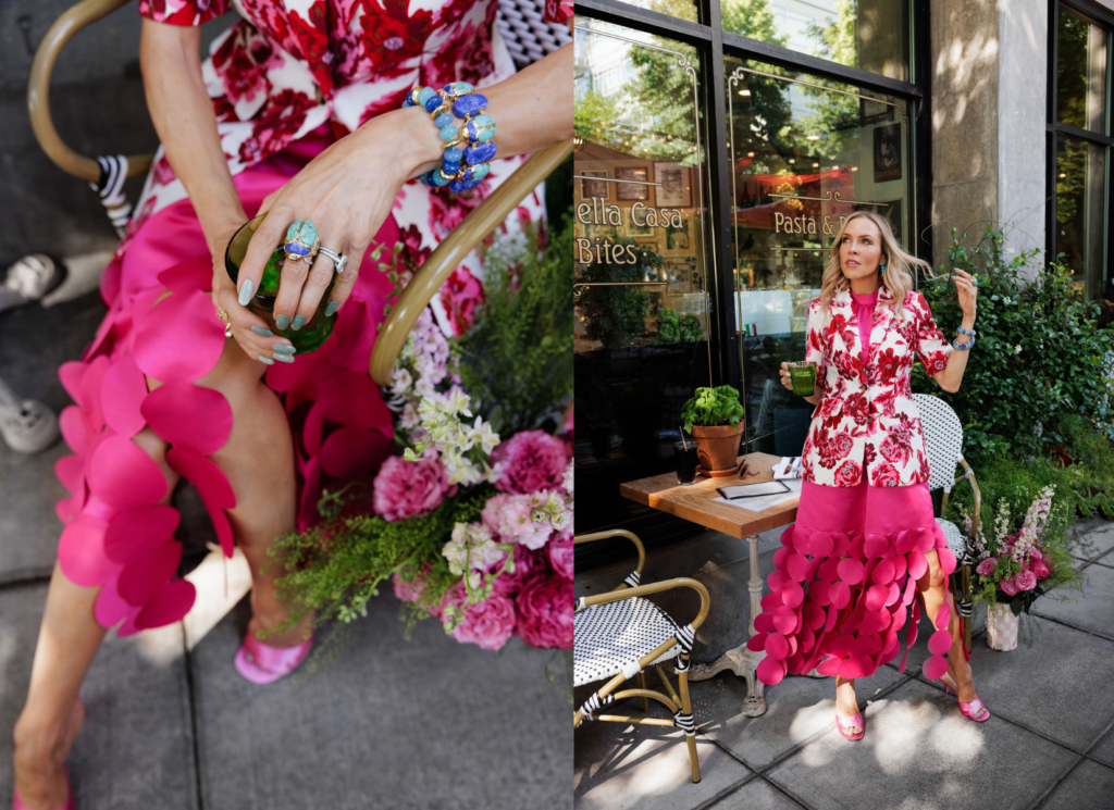Woman wearing a pink floral blazer and bright pink petal skirt while holding a bouquet of flowers outside a café.