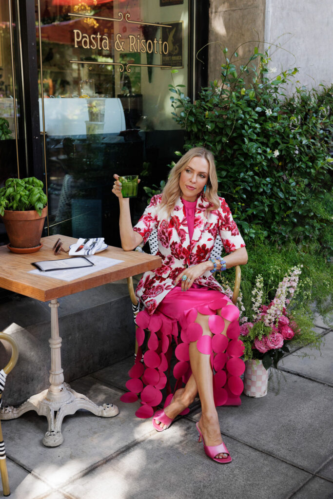 Woman wearing a pink floral blazer and bright pink petal skirt while holding a bouquet of flowers outside a café.