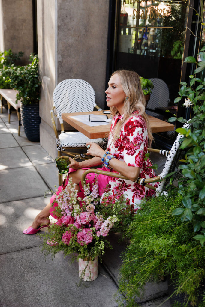 Woman wearing a pink floral blazer and bright pink petal skirt while holding a bouquet of flowers outside a café.