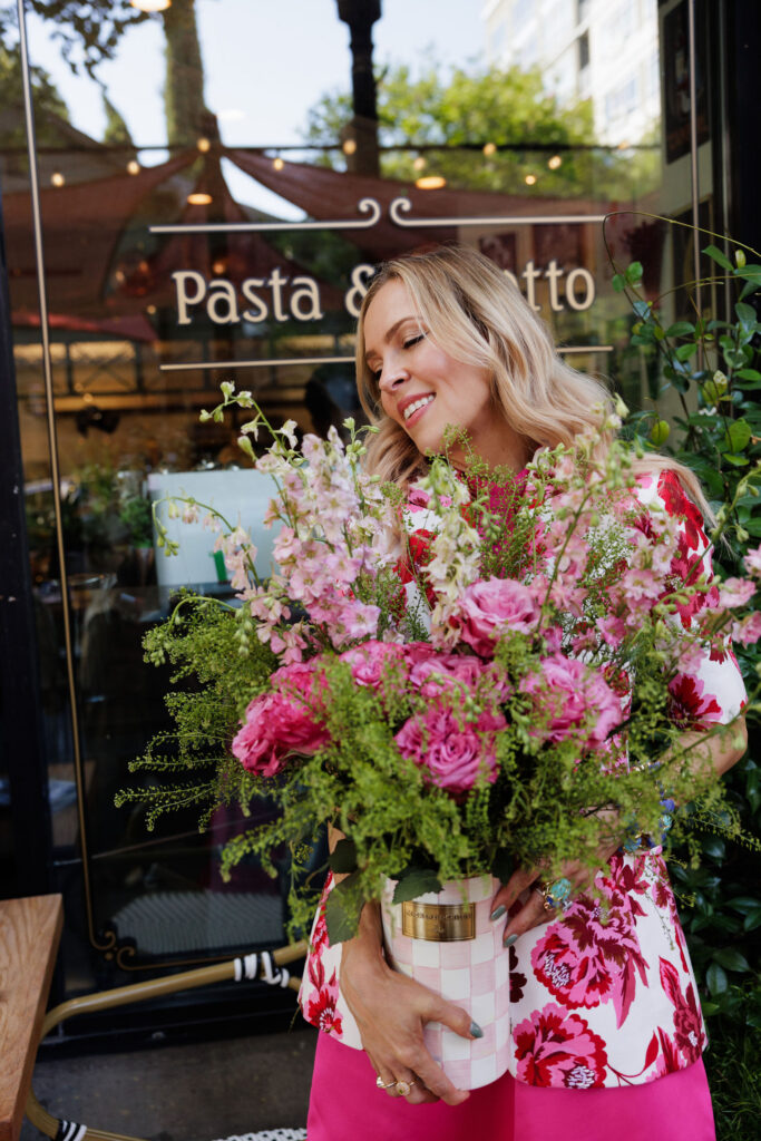 Woman wearing a pink floral blazer and bright pink petal skirt while holding a bouquet of flowers outside a café.