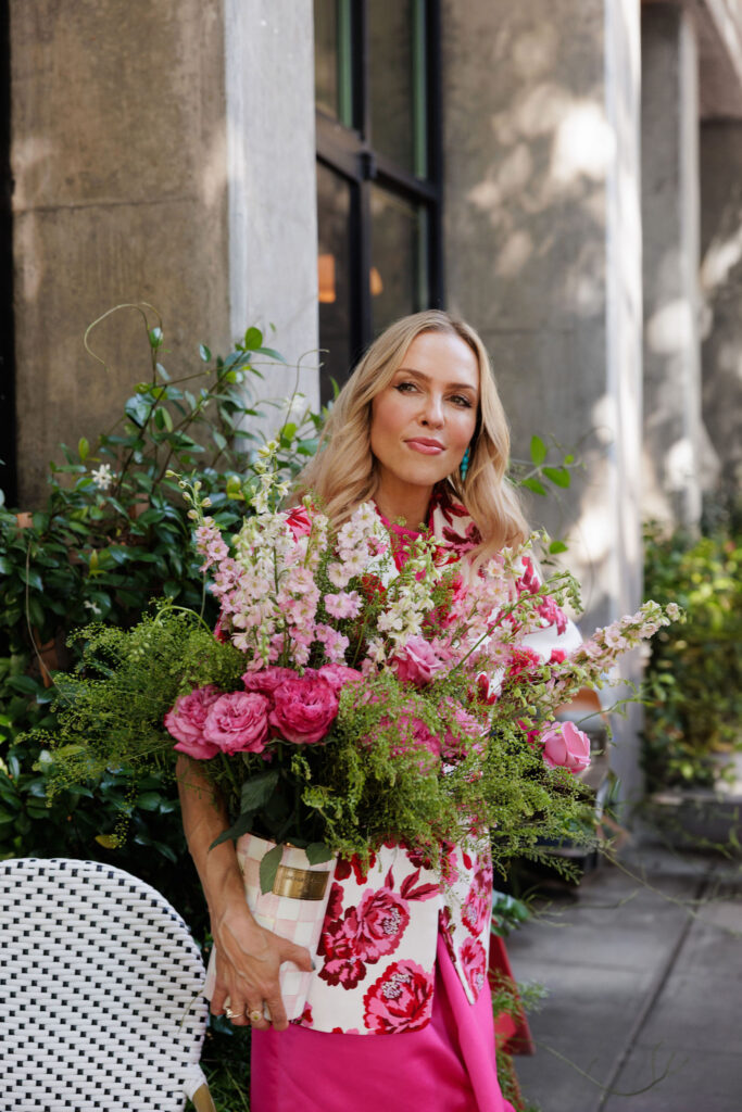 Woman wearing a pink floral blazer and bright pink petal skirt while holding a bouquet of flowers outside a café.