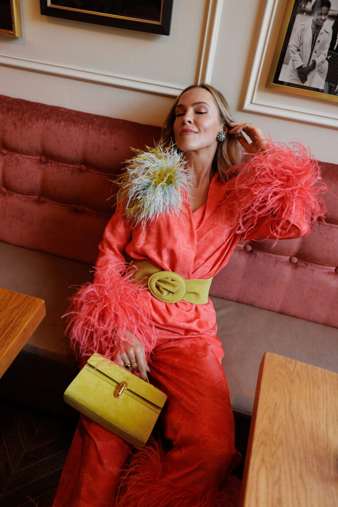 Coral satin suit styled with feather cuffs and a colorful feather brooch for a joyful statement look.