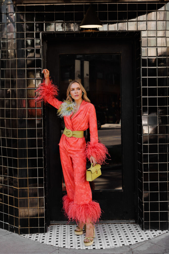 Coral satin suit styled with feather cuffs and a colorful feather brooch for a joyful statement look.
