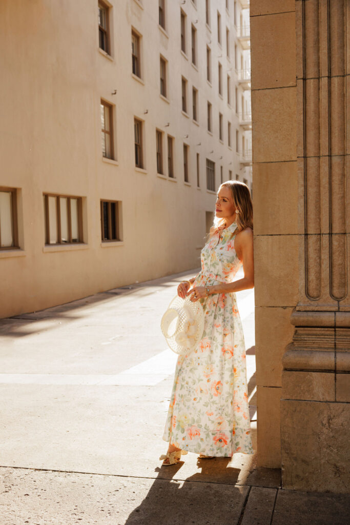 Woman wearing a pastel floral Needle & Thread maxi dress holding a straw hat in golden afternoon light