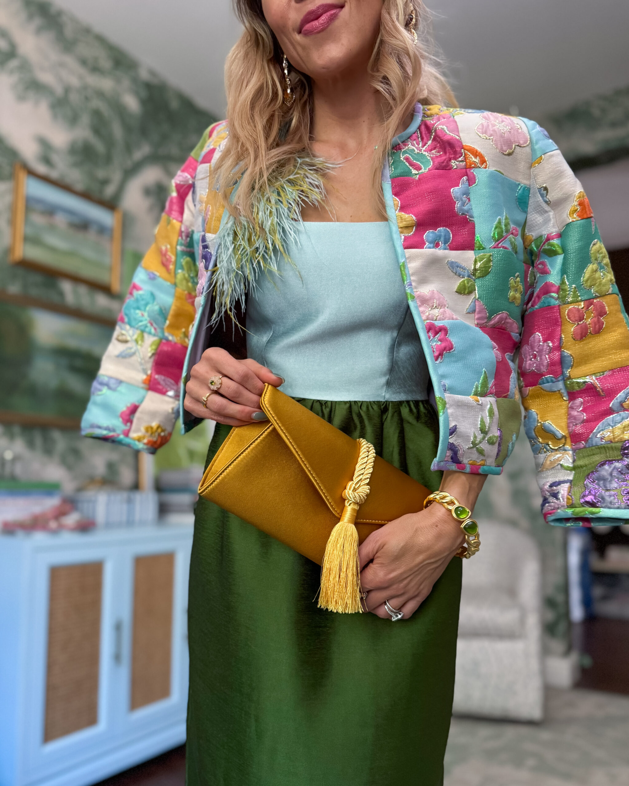 Woman wearing a pastel blue and olive green feather-trim colorblock dress styled with a colorful patchwork floral cropped jacket, mustard satin clutch, and green statement jewelry.
