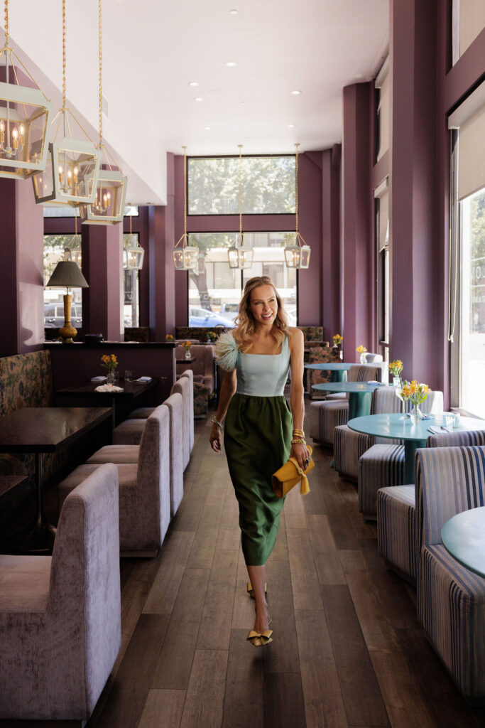 Woman wearing a color-block summer wedding guest dress while walking through a stylish restaurant with colorful accessories.