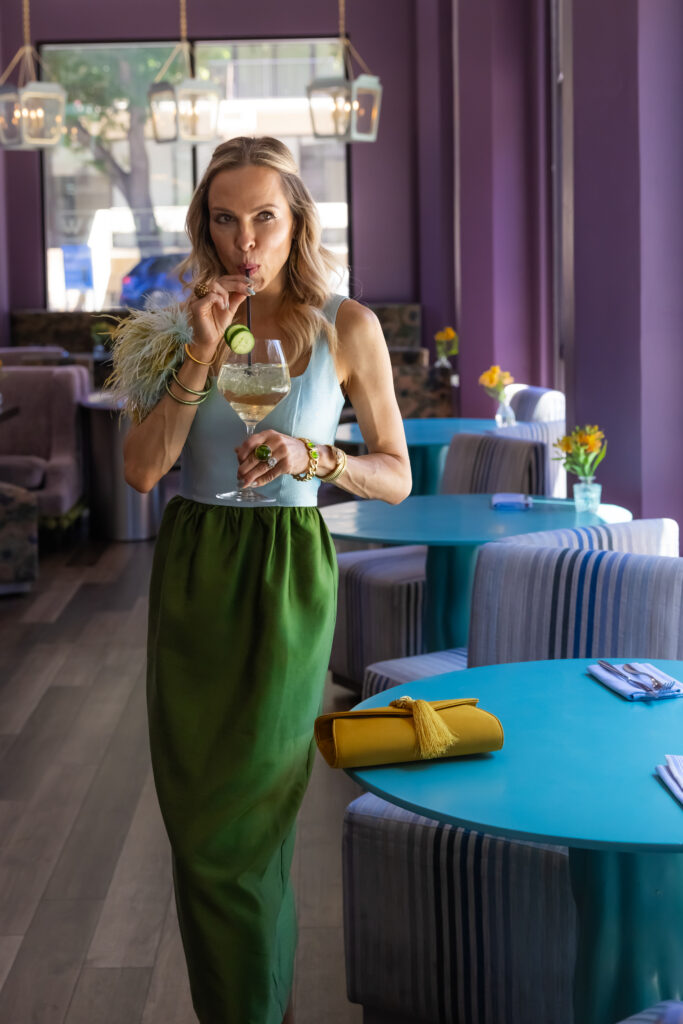Woman wearing a pastel blue and olive green feather-trim colorblock dress styled with a colorful patchwork floral cropped jacket, mustard satin clutch, and green statement jewelry.