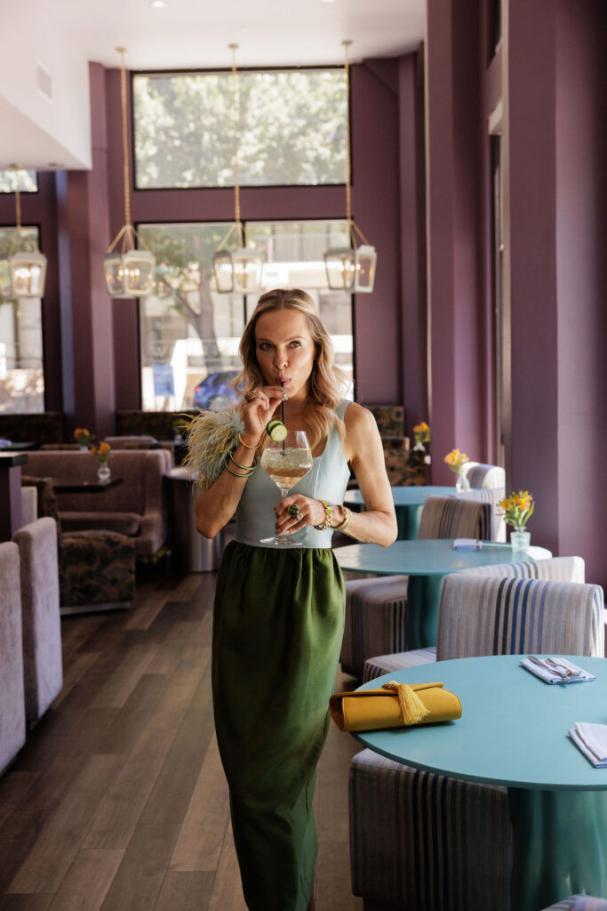 Woman wearing a color-block summer wedding guest dress while standing in a stylish restaurant sipping a drink with colorful accessories.