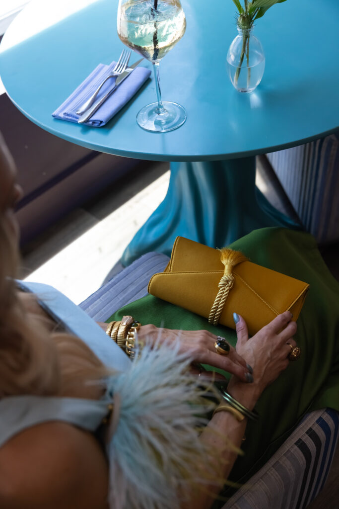 Woman wearing a pastel blue and olive green feather-trim colorblock dress styled with a colorful patchwork floral cropped jacket, mustard satin clutch, and green statement jewelry.