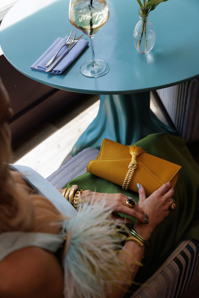 Woman wearing a blue and olive green color-block Tuckernuck dress with feather shoulder detail, yellow clutch and green statement jewelry.