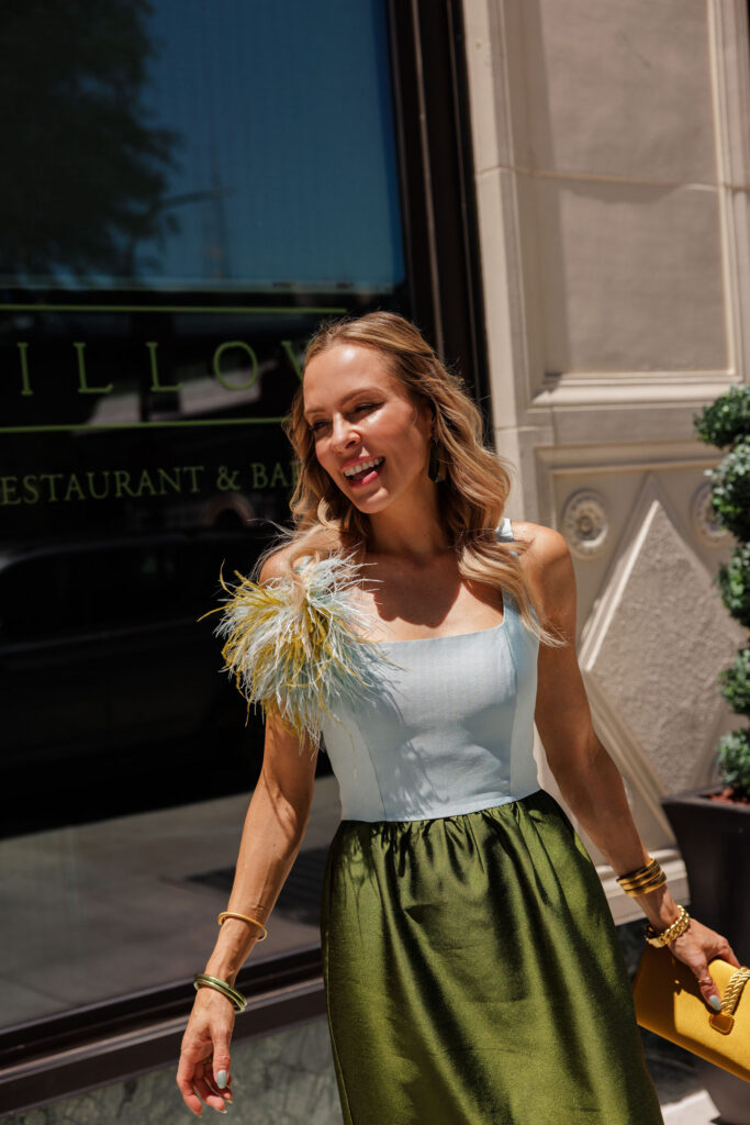 Woman wearing a blue and olive green color-block Tuckernuck dress with feather shoulder detail, yellow clutch and green statement jewelry.