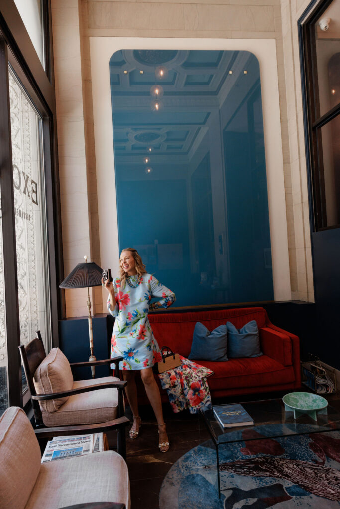 Woman wearing a light blue floral summer dress with coral red flowers and turquoise jewelry, showing how to style pops of red for summer fashion.